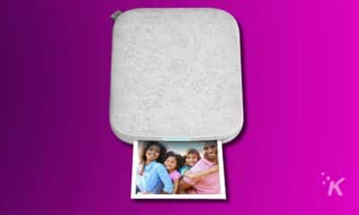 An HP Sprocket portable photo printer on a magenta background with a printed photo of a smiling family with two adults and two children.